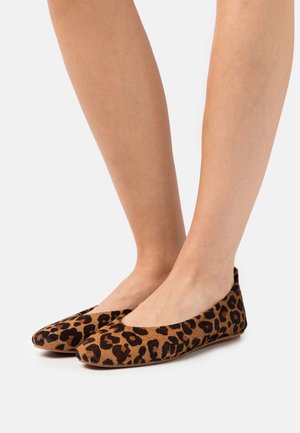 Een paar voeten in bruine en zwarte leopardprint balletschoenen tegen een effen witte achtergrond.