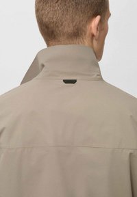 Beige waterdichte jas met een hoge kraag, voorzien van zwarte hardware aan de achterkant en een gladde textuur. Schoon ontwerp zonder zichtbare patronen.