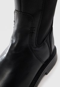 Gros plan sur une botte en cuir noir avec un panneau latéral élastique et des coutures détaillées sur la tige et la partie supérieure du pied.