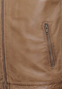 Ruskea nahkatakki, jossa on sileä pinta, hillitty kiilto ja sivuvetoketjullinen tasku, jossa on hopeinen varustus. Saumojen yksityiskohtia reunoilla.