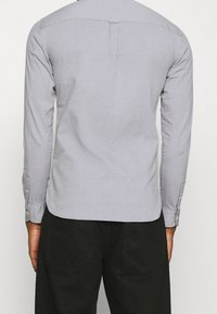 Camicia grigia chiaro a maniche lunghe realizzata in tessuto con una texture liscia. Presenta un orlo curvo, una piega sul retro e polsini con bottoni.