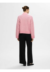 Un pull rose, court, en matière douce avec un col montant et des épaules tombantes ; porté avec un pantalon noir ample et des chaussures plates.