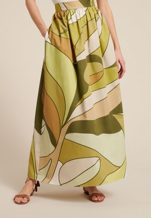 Kobieta ubrana w długą spódnicę z wysokim stanem, ozdobioną dużymi abstrakcyjnymi liśćmi w kolorach zielonym, beżowym i brązowym oraz brązowe sandały z odkrytymi palcami.