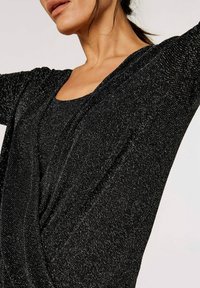 Haut en maille noire avec une finition texturée et un design drapé. Il présente un col large et des manches amples, mettant en avant le confort et la polyvalence.