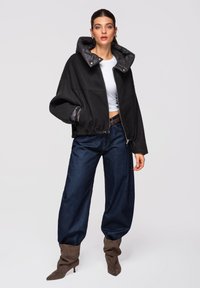 Femme portant une veste noire oversize avec un col matelassé, un crop top blanc, un jean large foncé et des bottes marron à talons souples, debout devant un fond uni.