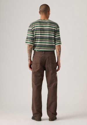 Person mit Tätowierungen auf Armen und Hals, trägt ein grün gestreiftes Kurzarmhemd und weite braune Hosen, steht mit dem Rücken zum Betrachter vor einem einfarbigen Hintergrund.