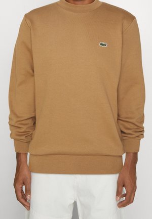 Mężczyzna ubrany w camelowy brązowy sweter crewneck z małym zielonym logo krokodyla na piersi oraz białe spodnie.