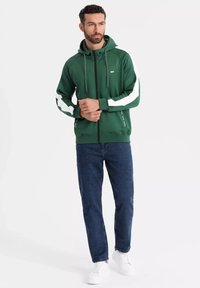 Felpa verde con zip e strisce bianche ai lati, cappuccio con cordino e tasche anteriori, abbinata a jeans blu scuri e sneakers bianche.
