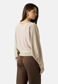 Maglione beige con maniche larghe e orlo a coste, caratterizzato da un dettaglio a catena dorata sul retro. Abbinato a pantaloni marroni.