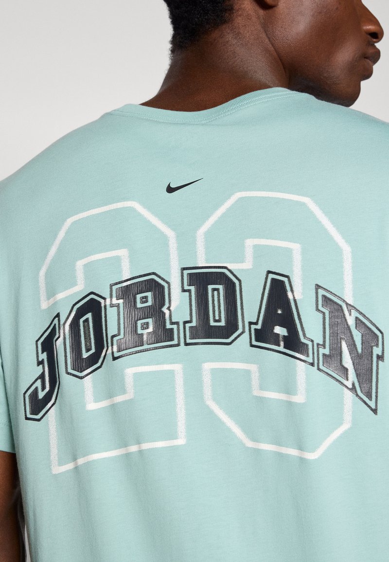 Camiseta de color azul claro con "JORDAN" y "23" impresos en negro y blanco en la parte trasera. Presenta un pequeño logo negro de Nike en la parte superior de la espalda.