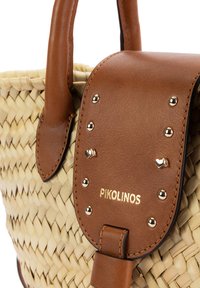 Bolso de paja tejido con solapa de cuero marrón, tachuelas doradas, pequeños detalles en forma de corazón y logo Pikolinos en relieve en el cierre de la solapa.