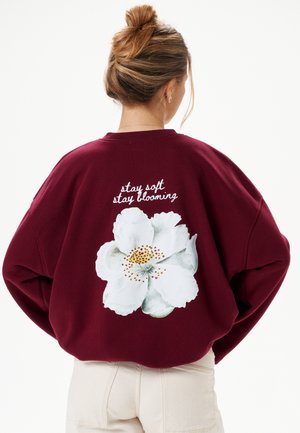 Vrouw met bruin haar in een knot draagt een bordeauxrode trui met een grote witte bloem en de tekst "stay soft stay blooming" op de rug.