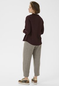 Camicia bordeaux a maniche lunghe con colletto a frill, abbinata a pantaloni tapered grigi chiari e sneakers marroni con dettagli a texture mista.
