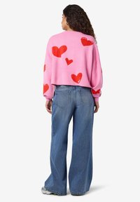 Ροζ cropped πουλόβερ με κόκκινα μοτίβα καρδιάς, συνδυασμένο με φαρδιά μπλε τζιν παντελόνια. Το ύφασμα φαίνεται μαλακό με άνετη εφαρμογή.