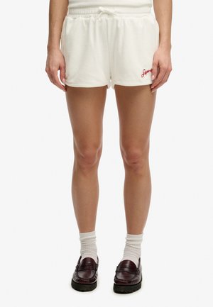 Kvinde iført hvide shorts med snøre og rød logo, hvide sokker og mørkebrune loafers, stående mod en hvid baggrund.