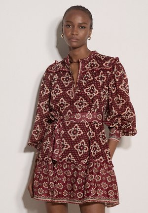 Donna con i capelli intrecciati che indossa un vestito bordeaux con motivo floreale, maniche a sbuffo e cintura annodata in vita, in piedi su uno sfondo semplice.