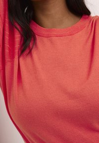 Gros plan d'une personne portant un pull en maille rose corail avec un col rond et de longs cheveux noirs partiellement visibles.