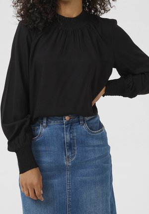 Femme portant un chemisier noir à manches longues avec poignets et encolure smockés, associé à une jupe en denim bleu taille haute.