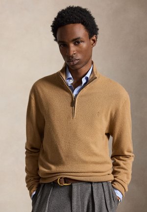 Ung mand med krøllet hår, iført en tan kvart-lynlås sweater over en lyseblå skjorte, grå plisserede bukser og et bælte, stående med hænderne i lommerne.