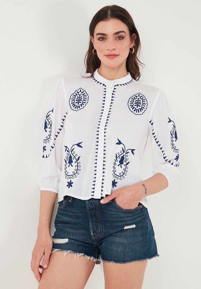Witte blouse met blauwe geborduurde patronen, voorzien van een staande kraag, knopen aan de voorkant en driekwart mouwen, gecombineerd met denim shorts.