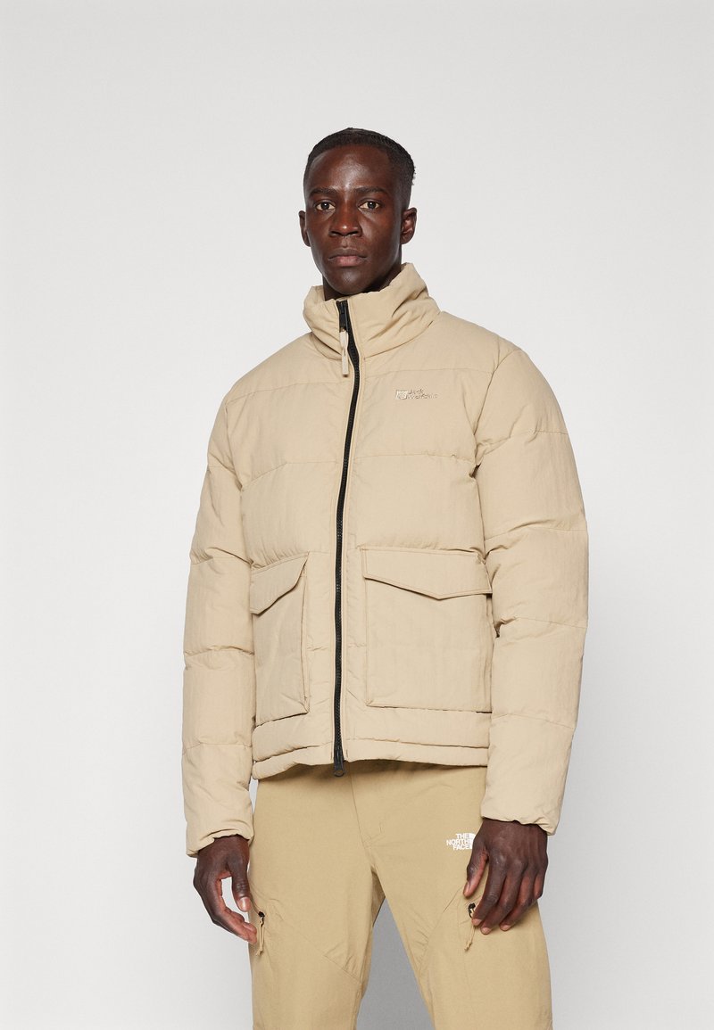 Beige pufferjack met een hoge kraag, voorrits en twee grote borstzakken. Zachte textuur en gequilt ontwerp voor warmte.