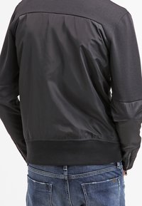 Schwarze Bomberjacke mit einer glatten Satin-Oberfläche und gerippten Bündchen. Beinhaltet ein kontrastierendes mattes Material auf der oberen Rückseite und den Schultern.