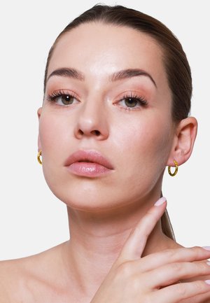 Boucles d'oreilles en or avec une finition texturée, portées par une personne avec un maquillage naturel et un arrière-plan neutre, mettant en valeur les traits du visage et la position des mains.