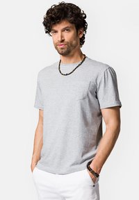 Szara bawełniana koszulka z okrągłym dekoltem i krótkimi rękawami, z kieszonką na lewej piersi. Noszona z kolczykami z ciemnych koralików, model na neutralnym tle.