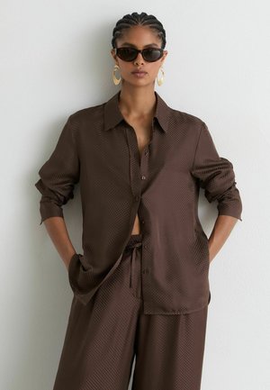 Vrouw met gevlochten haar die donkere zonnebril, grote gouden oorbellen en een gestructureerd donkerbruin shirt- en broekenset draagt tegen een witte muur.