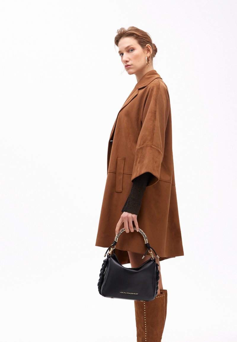 Cappotto oversized in suede marrone con maniche larghe, indossato sopra un maglione in maglia scura. Il modello tiene in mano una borsa in pelle nera con manico intrecciato e dettagli dorati.