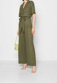 Olivgrön jumpsuit med korta ärmar, knytmidja, vida ben, två framfickor, kombinerad med en liten ljusgrön väska och klackar.