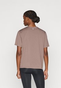 Καφέ κοντομάνικο T-shirt με στρογγυλή λαιμόκοψη, με υφή υφάσματος. Λογότυπο "S" στο πάνω μέρος της πλάτης. Συνδυασμένο με μαύρο κολάν.