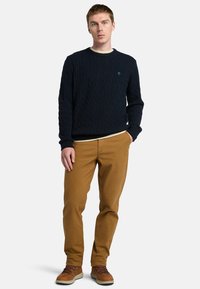 Maglione navy a trecce con scollo rotondo, abbinato a chinos beige e stivali marroni con lacci, in piedi davanti a uno sfondo bianco.