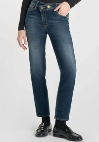 Jean en denim bleu foncé avec une coupe droite, décoloration légère, coutures contrastées, fermeture à deux boutons et poches. Porté avec des mocassins noirs.