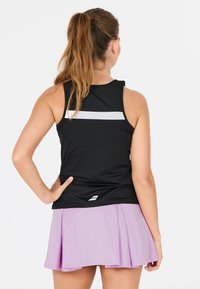 Donna che indossa un top sportivo senza maniche nero con una striscia bianca e una gonna plissée lavanda, in piedi con una mano sul fianco.