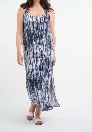 Kobieta ubrana w bezrękawową, granatowo-białą sukienkę maxi tie-dye z bocznym rozcięciem, zestawioną z białymi, wsuwanymi sandałami, stojąca na białym tle.