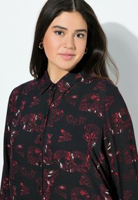 Maglietta nera con un design a colletto, caratterizzata da un motivo astratto rosso. Realizzata in un tessuto con una texture liscia e chiusura a bottoni.