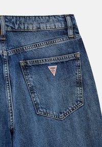 Dunkelblaue Denim-Jeans mit einer dreieckigen Gesäßtasche, die mit einem roten und weißen "GUESS"-Logopatch und kontrastierender goldener Naht verziert ist.