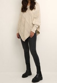 Beige gebreid poncho-trui met chevronpatronen; losse pasvorm; lange mouwen; gecombineerd met zwarte skinny jeans en zwarte haklaarzen.