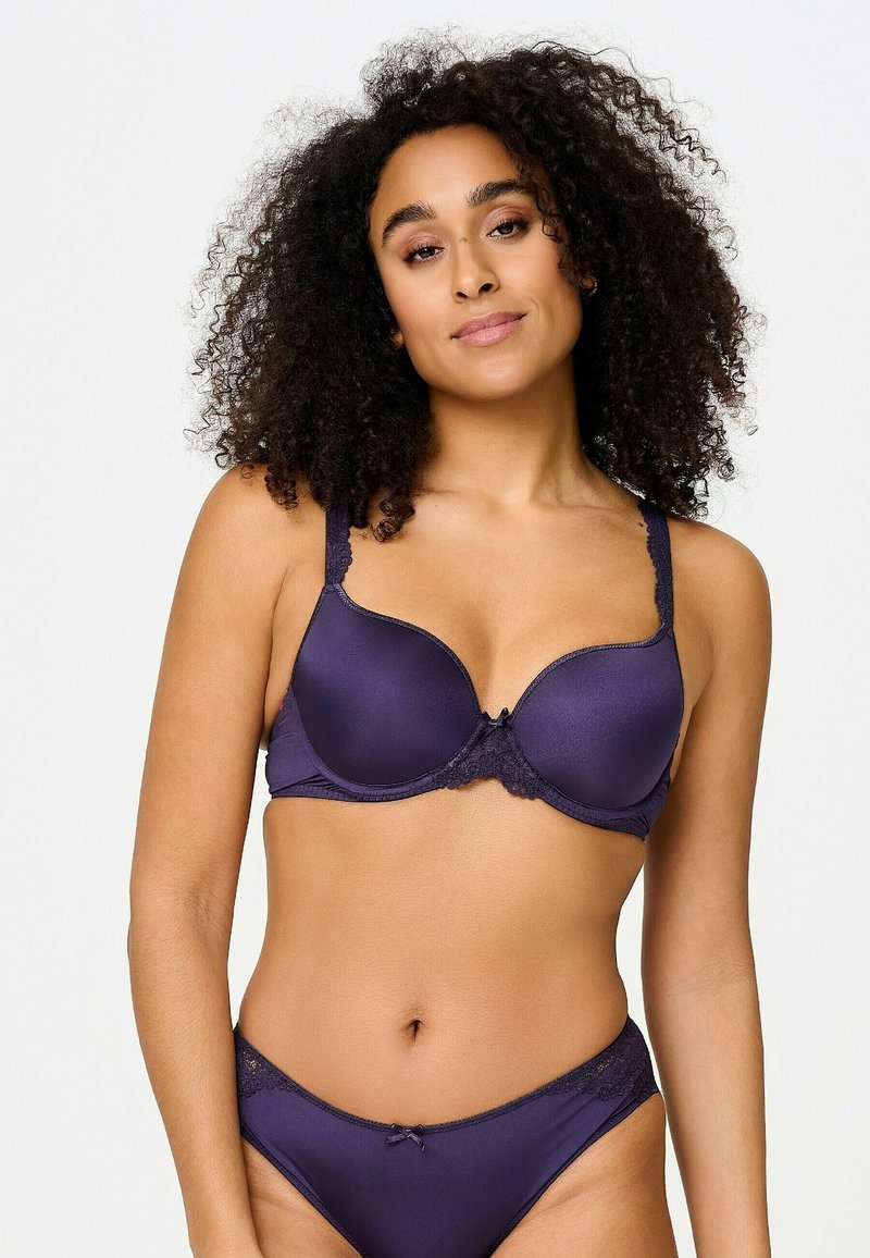 Femme aux cheveux bouclés portant un soutien-gorge et une culotte assortis violet foncé avec des détails en dentelle, debout devant un fond clair uni.