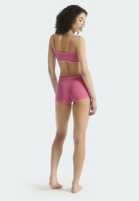 Femme aux cheveux bouclés se tient pieds nus de dos, portant un soutien-gorge de sport rose et un short assorti, devant un fond clair.