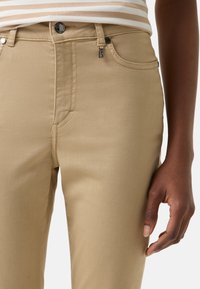 Beige katoenen broek met een smalle pasvorm, voorzakken en een ritssluiting. Metalen knoop en labeldetail op de tailleband. Soepele textuur.