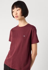 Young person with short black hair wears a plain maroon t-shirt with a small logo on the chest, standing against a white background.
