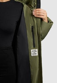 Olive krāsas izolēta jaka ar melnu iekšpusi, kurai ir rāvējslēdzēja kabata un uzlīme ar tekstu "Silta apskāviens, kas izgatavots no auduma."