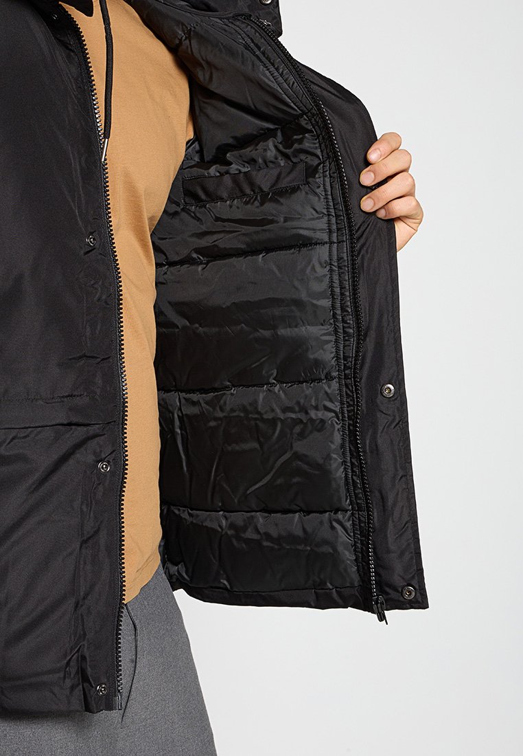 Veste noire isolée avec un motif matelassé, dotée d'une fermeture éclair et d'une poche intérieure, présentée avec la doublure intérieure visible.