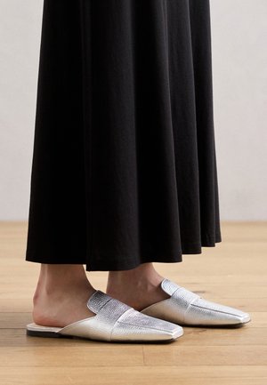 Sølvskinn slip-on sko med teksturert overflate, spiss design og åpen bak, kombinert med et langt svart skjørt.