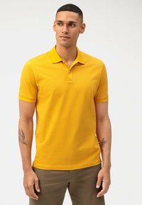 Gele polo met een klassieke kraag, korte mouwen en een drieknoopsluiting. Gladde textuur; gedragen met lichtbruine broek.