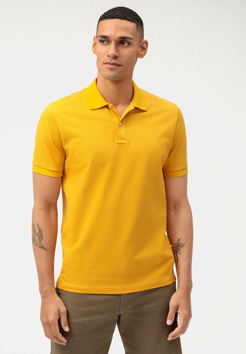 Gele polo met een klassieke kraag, korte mouwen en een drieknoopsluiting. Gladde textuur; gedragen met lichtbruine broek.