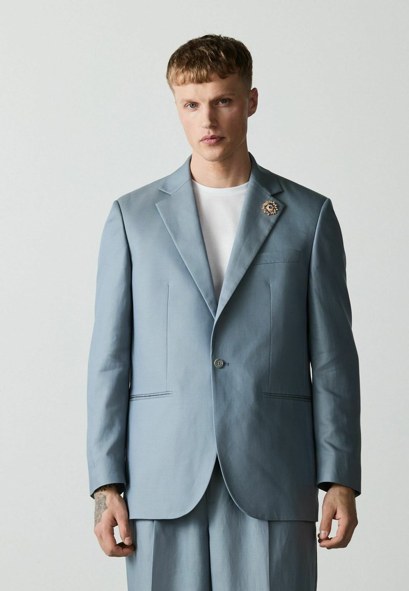 Homme portant un costume bleu clair avec une chemise blanche et une broche décorative, debout devant un fond gris uni, faisant face vers l'avant.