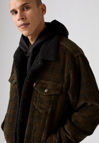 Brown corduroy jacket with a black, fuzzy collar; features two chest pockets and button details, layered over a black hooded sweatshirt.